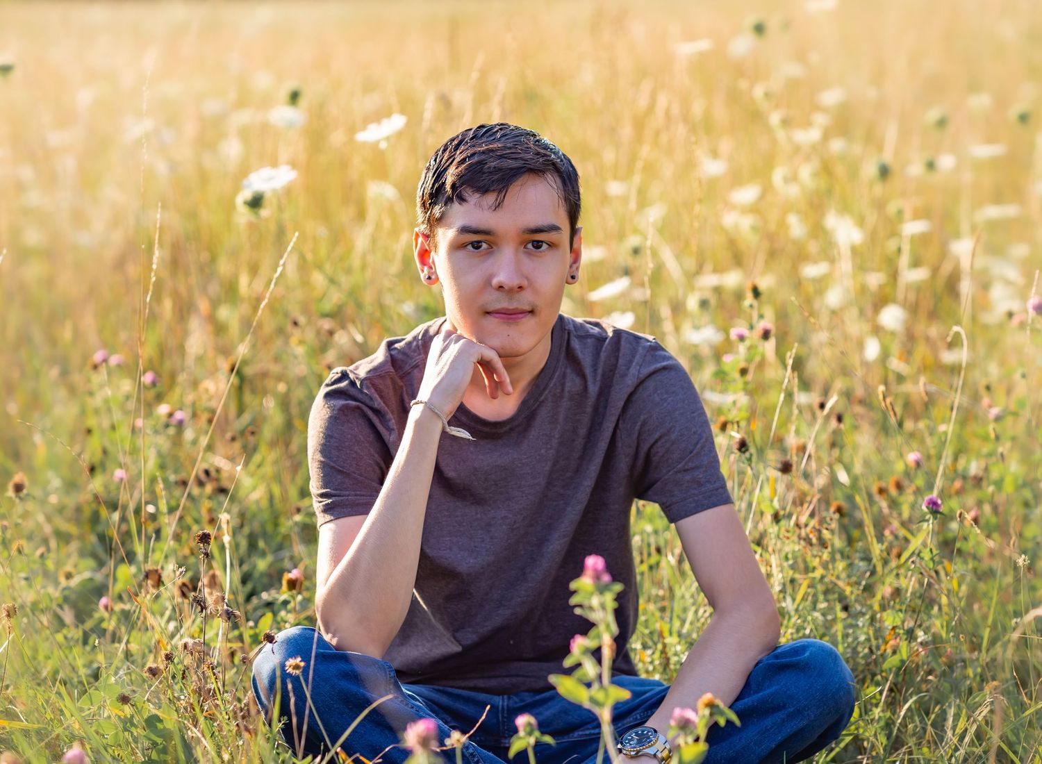 Young man sitting cross-legged in a sunlit wildflower meadow, resting chin on hand in a thoughtful pose.
