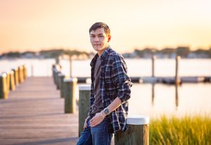 Young man in plaid shirt leaning on dock post at golden hour waterfront sunset backdrop.