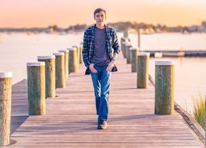 Young man in plaid shirt and jeans walking on a wooden pier at sunset with calm water in the background.