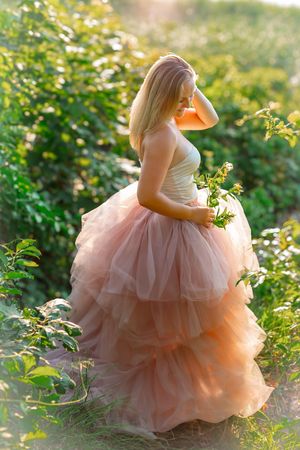 Blonde woman in a layered pink tulle ball gown standing in a sunlit green garden holding wildflowers.