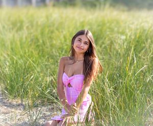 Young woman in pink strapless dress sitting among tall green grass in a sunny outdoor field setting.