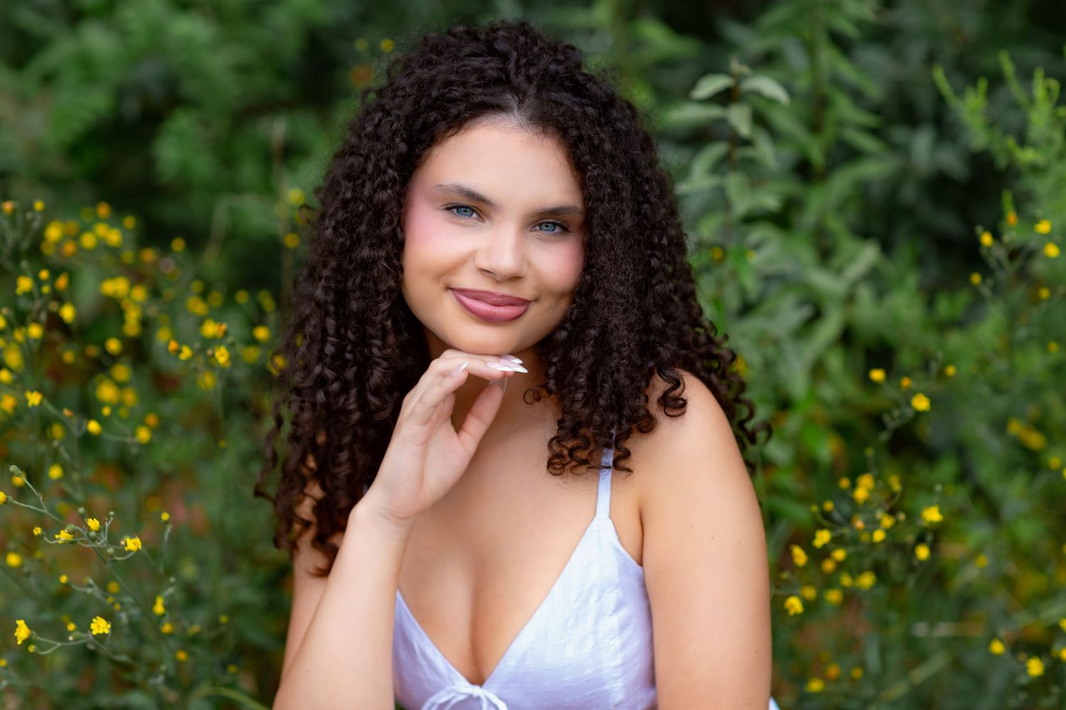 Young woman with curly hair smiling outdoors, wearing a white top, surrounded by lush green foliage and yellow flowers.