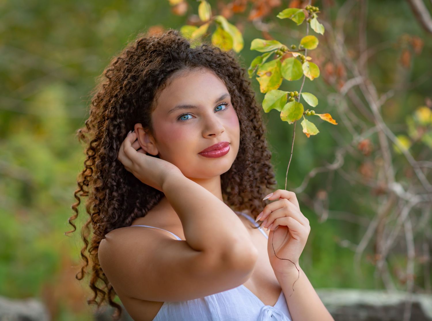 Young woman with curly brown hair and blue eyes holds a branch outdoors in a lush green nature setting.