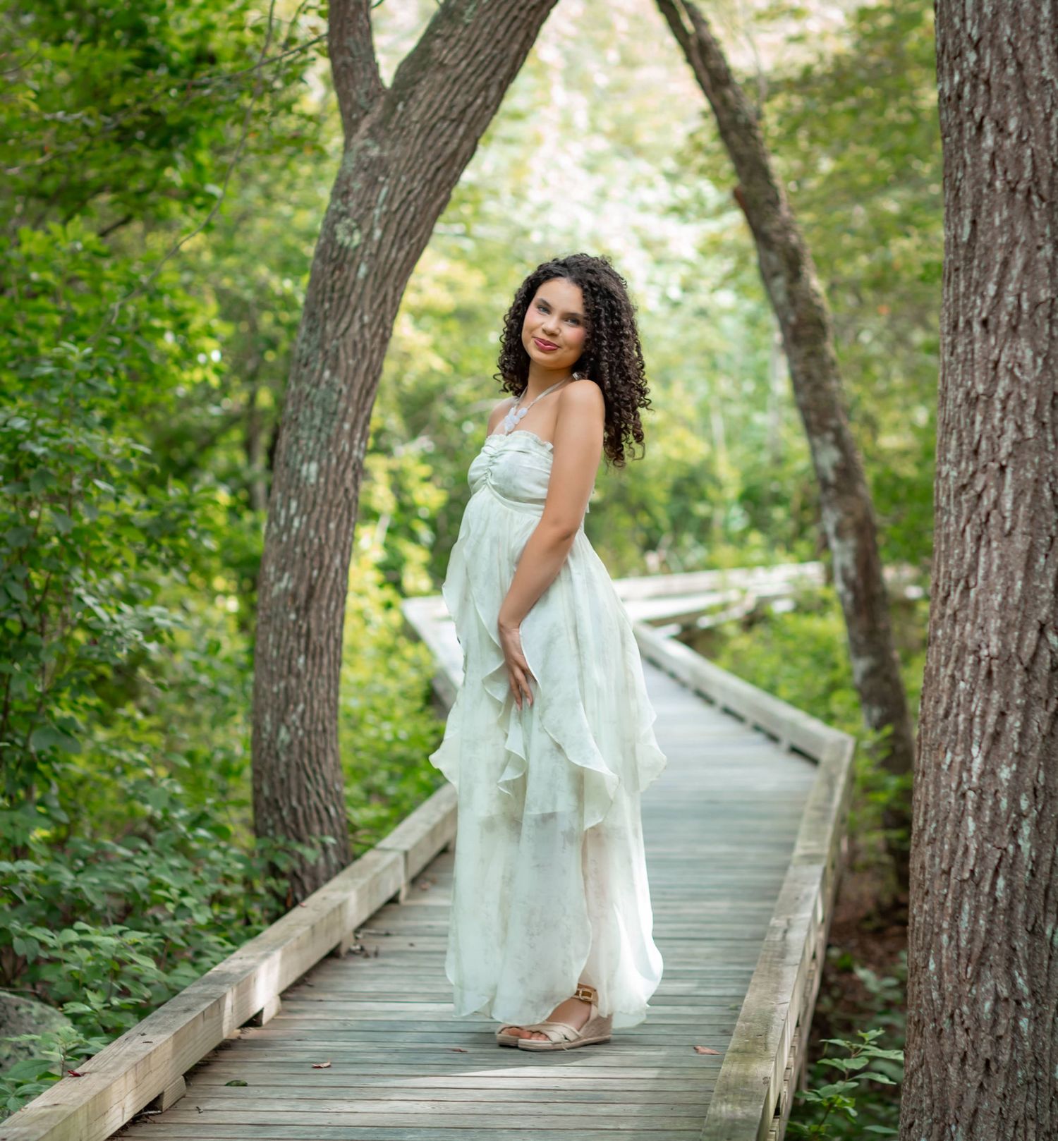 A young woman in a flowing white dress poses gracefully on a wooden boardwalk surrounded by lush green trees.