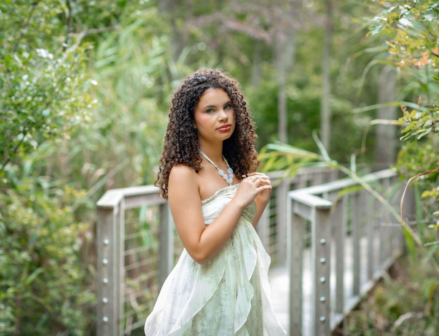 Young woman with curly hair in a white dress poses gracefully on a wooden bridge surrounded by lush green foliage.