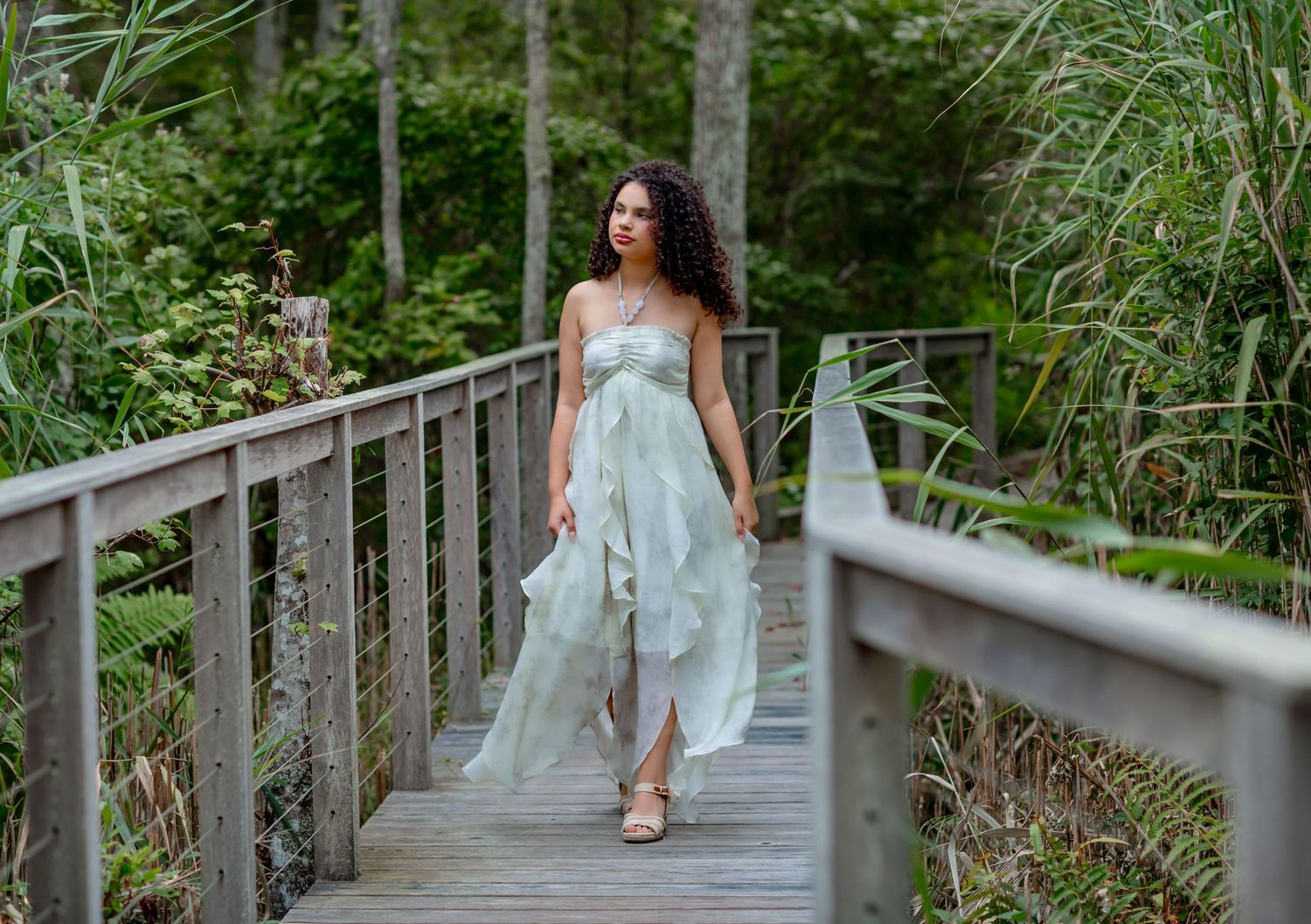 Woman in mint green dress walking on wooden boardwalk surrounded by lush green forest.