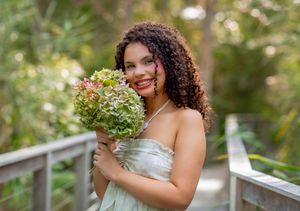 Smiling bride with curly hair holding green floral bouquet on wooden bridge surrounded by lush tropical greenery.