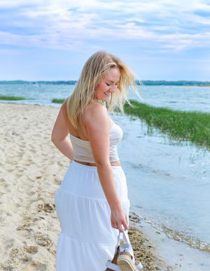 Blonde woman in white crop top and maxi skirt smiling on a sandy beach near calm water on a cloudy day.