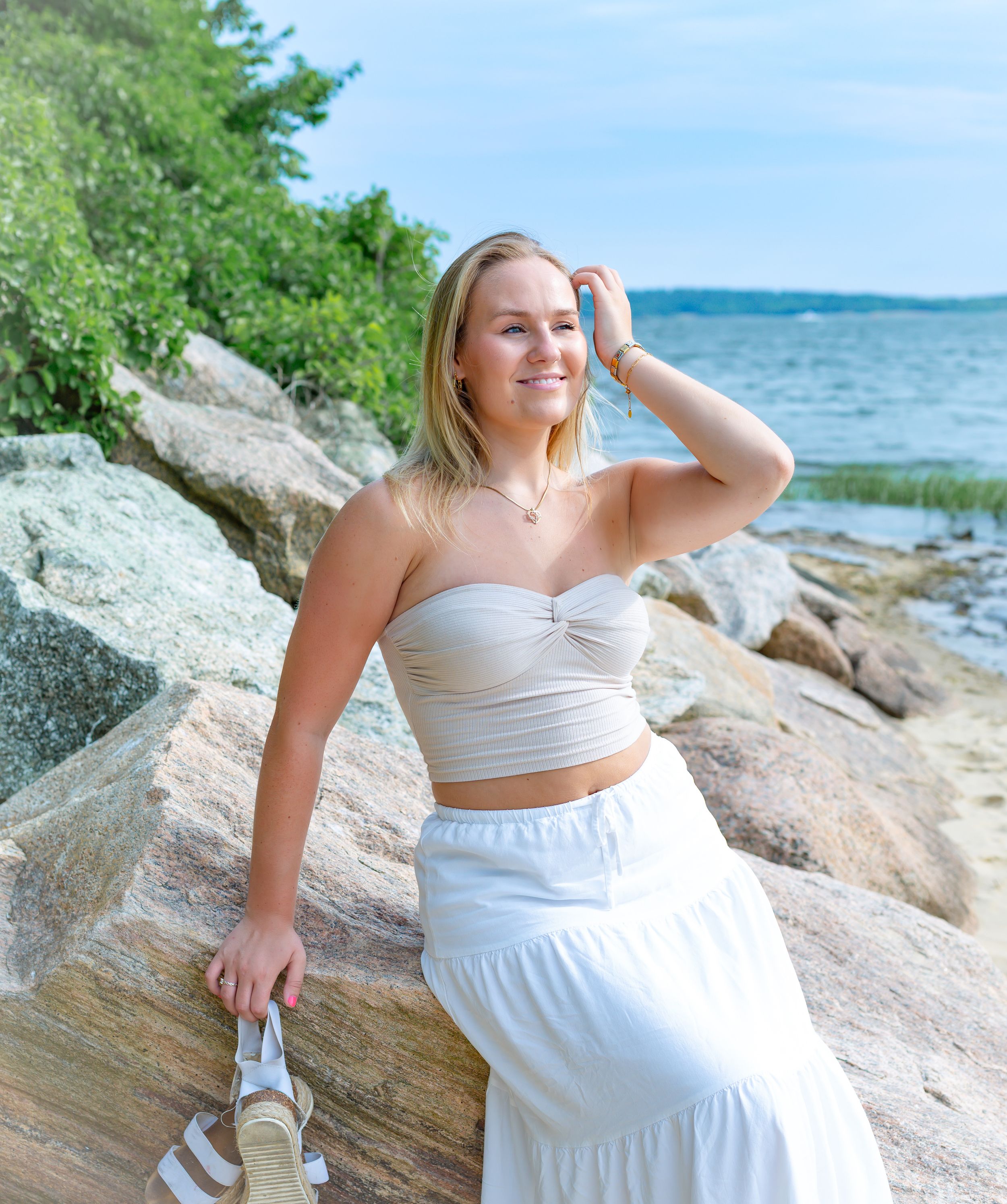 Smiling blonde woman in white crop top and maxi skirt posing on rocky shoreline with ocean views.