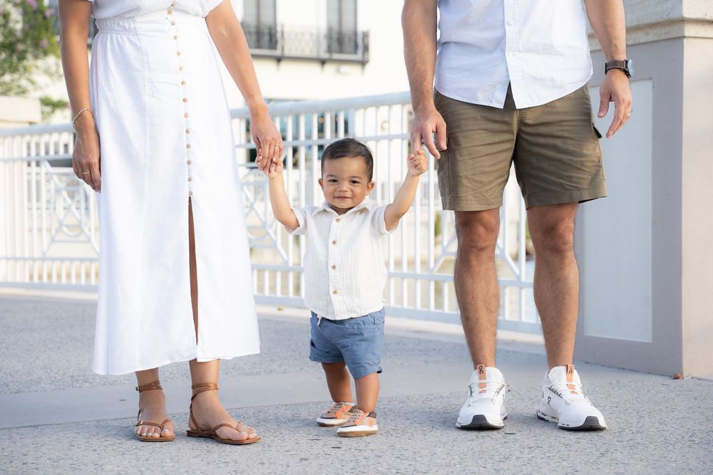 A toddler boy in white shirt and denim shorts holds hands with parents outdoors in a bright, sunny setting.