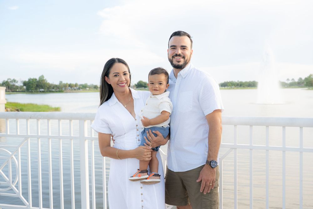 A young Hispanic family poses on a waterfront bridge, parents in white holding their baby boy on a sunny day.