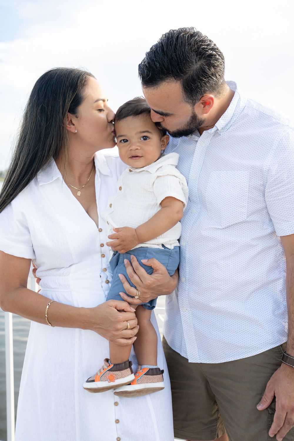A loving couple in white outfits kisses their baby boy outdoors against a bright sky backdrop.