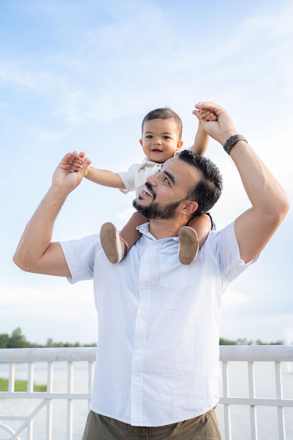 Father holding smiling baby on his shoulders outdoors against a blue sky on a sunny day.