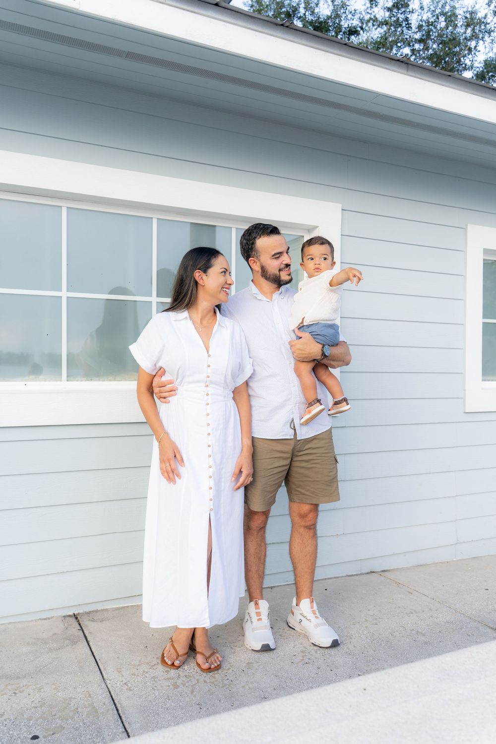 A young family of three poses outside a light blue house, with parents in white outfits and dad holding their toddler.