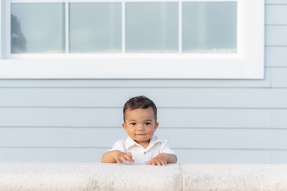 Adorable baby boy in white outfit peeking over a white ledge in front of a light blue house exterior.