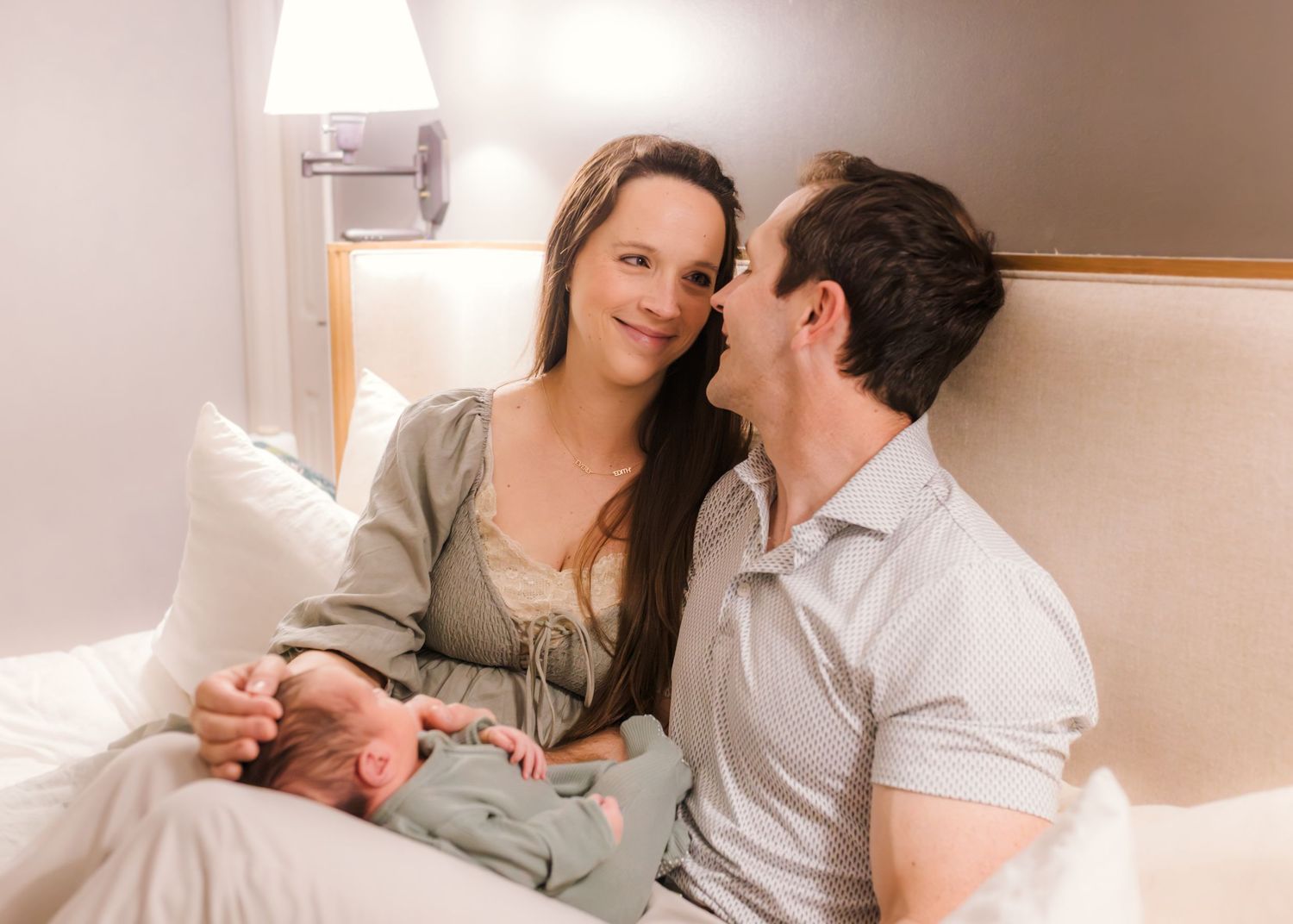 New parents sitting on a bed smiling at each other while lovingly holding their newborn baby.