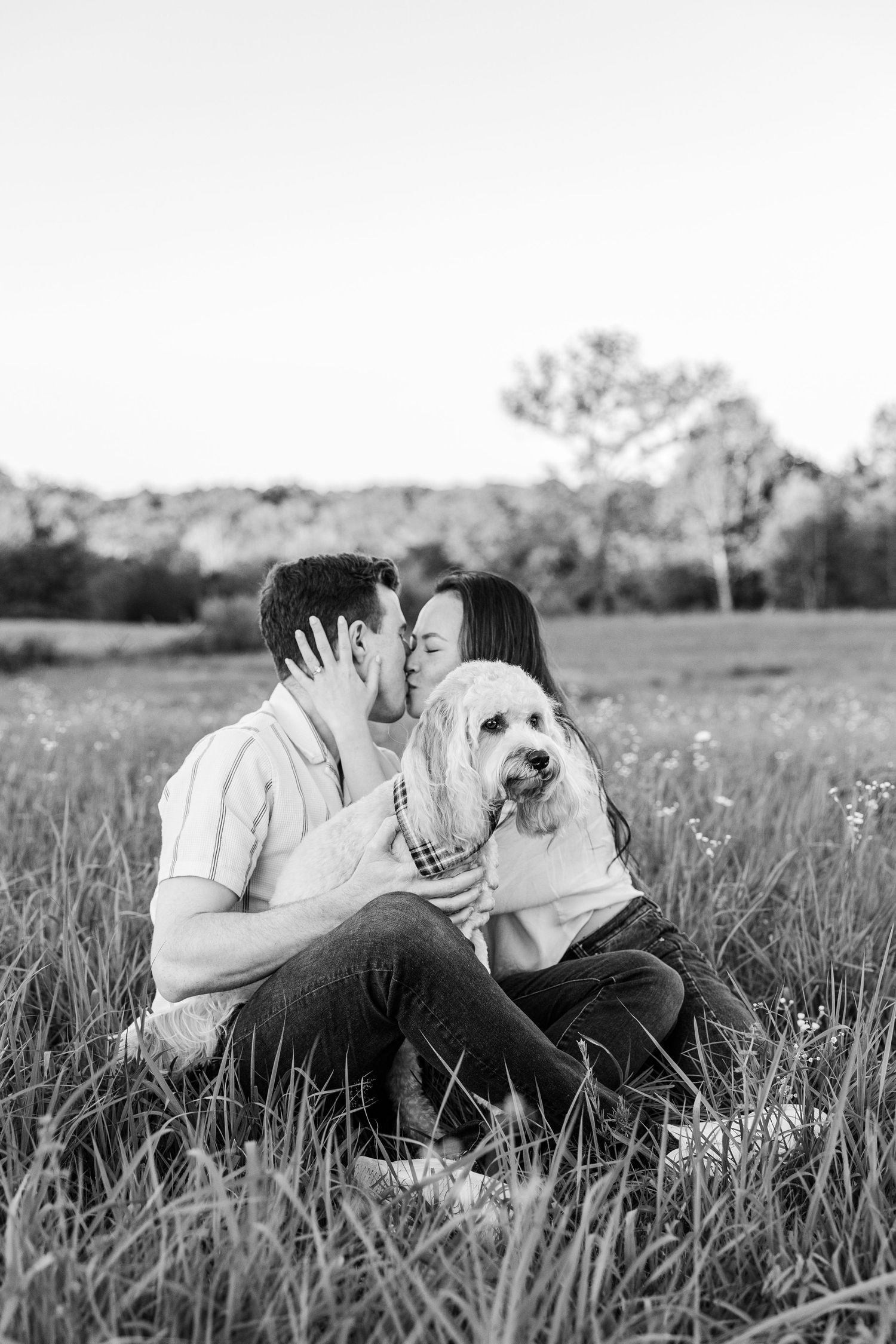 Black and white photo of a couple sitting in a field, sharing a kiss while holding their fluffy white dog.