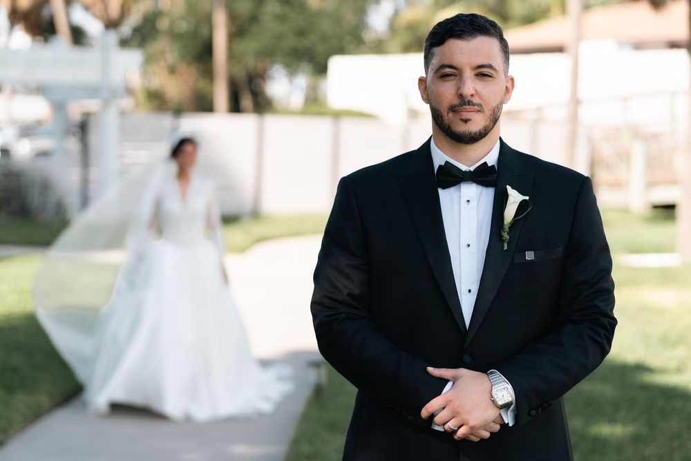 Groom in black tuxedo with bow tie stands confidently outdoors, bride in white wedding gown with veil in background.