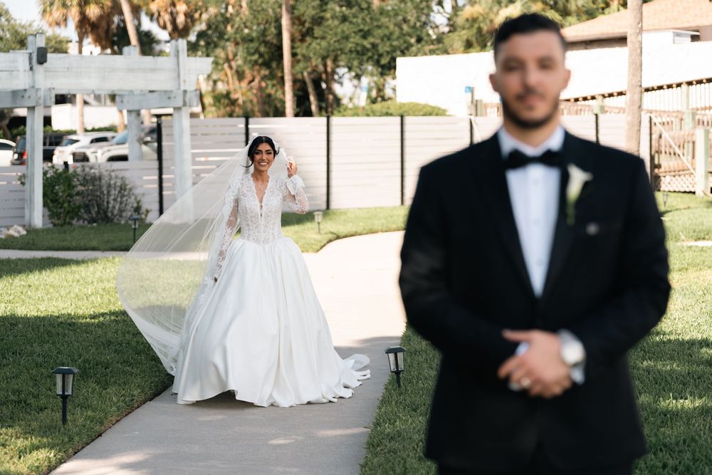 Bride in white gown waves approaching groom in black tuxedo during outdoor wedding first look ceremony.