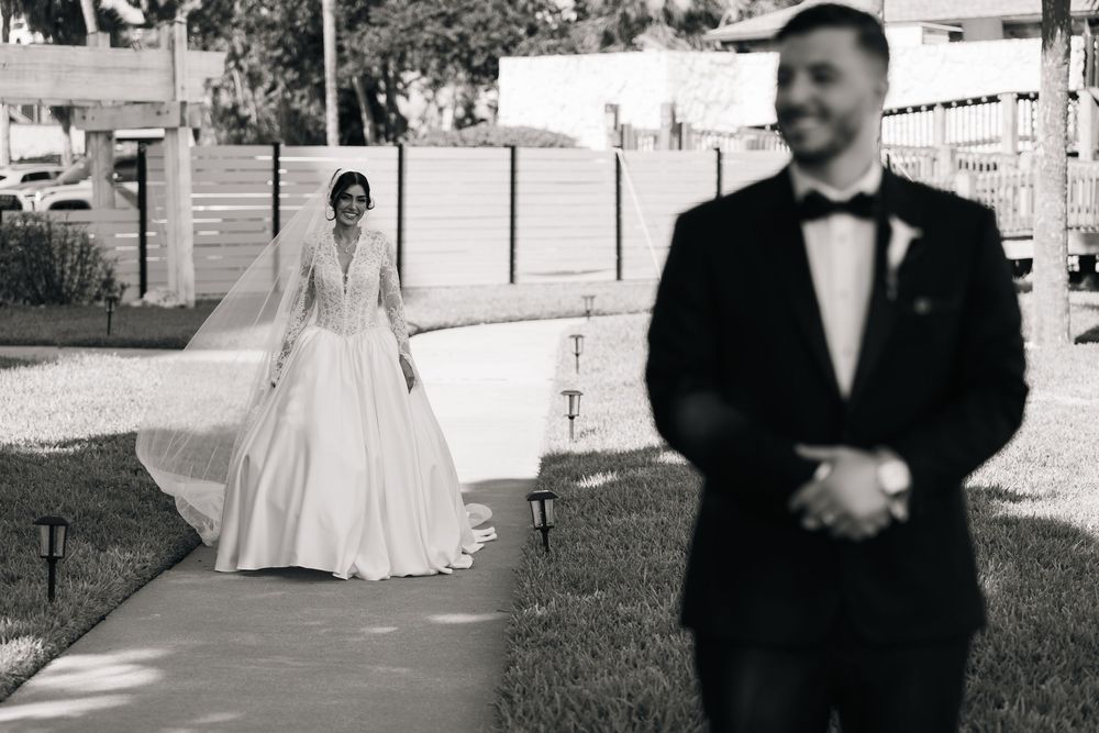 Black and white photo of a bride in a flowing gown and veil walking toward a groom in a tuxedo outdoors.