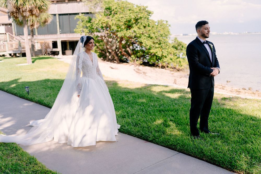 Bride in elegant long-sleeve ball gown with veil stands behind groom in black tuxedo for first look by waterfront.