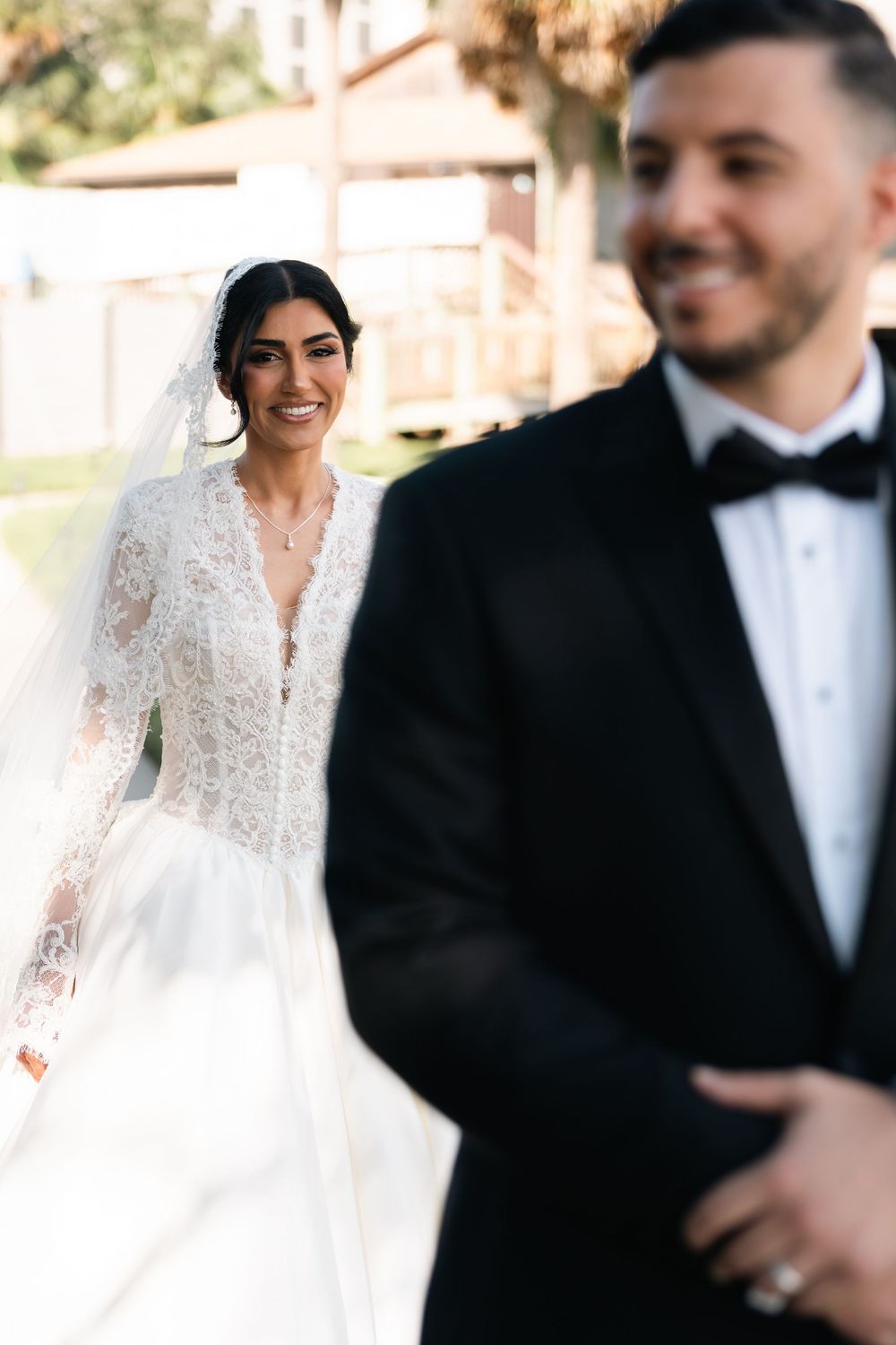 Smiling bride in lace long-sleeve wedding gown walks toward blurred groom in black tuxedo outdoors.
