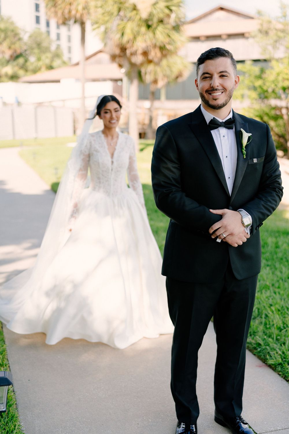 Groom in black tuxedo smiles outdoors as bride in lace wedding gown approaches on a sunny day.
