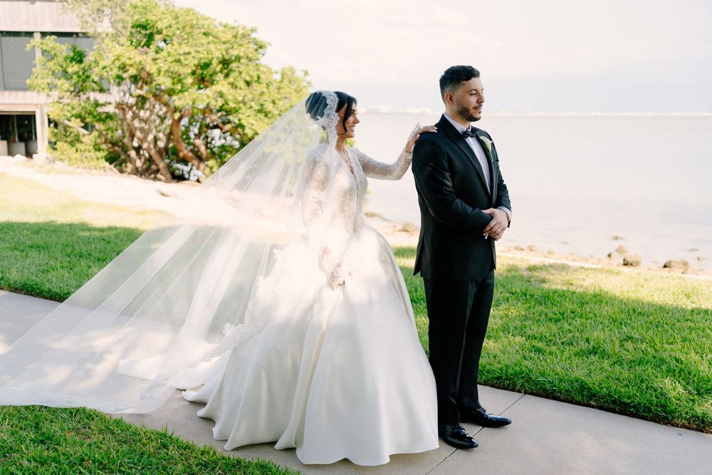 Bride in flowing white ball gown and veil stands with groom in black tuxedo outdoors near waterfront.