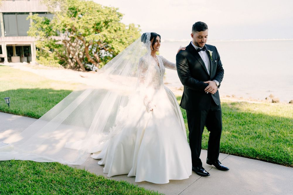 Bride in a ballgown with flowing veil stands beside groom in black tuxedo outdoors near waterfront.