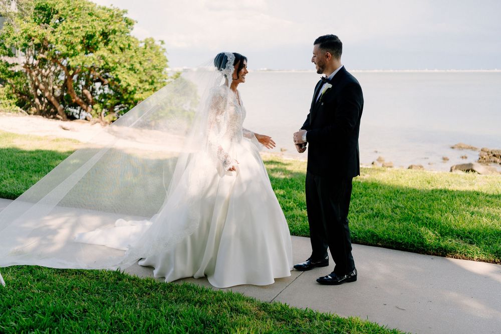 Bride and groom share first look moment by waterfront, veil billowing in wind, both in formal attire.