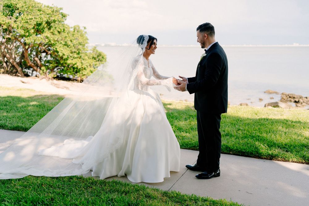 Bride in elegant white ball gown with long veil holds grooms hands during outdoor first look by waterfront.