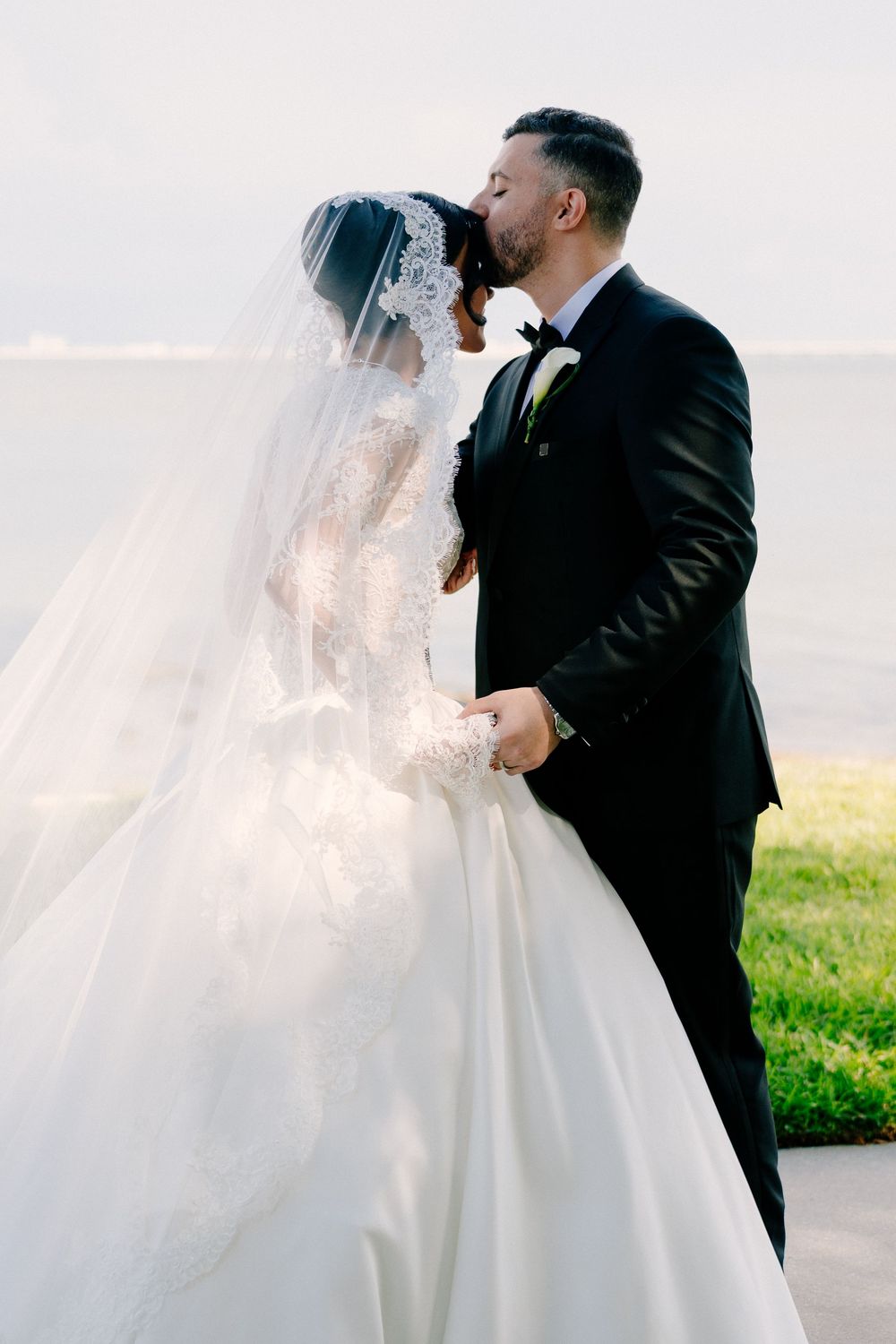 Bride and groom sharing a romantic kiss outdoors, bride wearing lace wedding gown with veil, groom in black tuxedo.