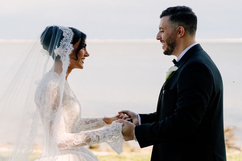 Bride and groom exchanging vows outdoors by the water, she in lace long-sleeve gown with veil, he in black tuxedo.