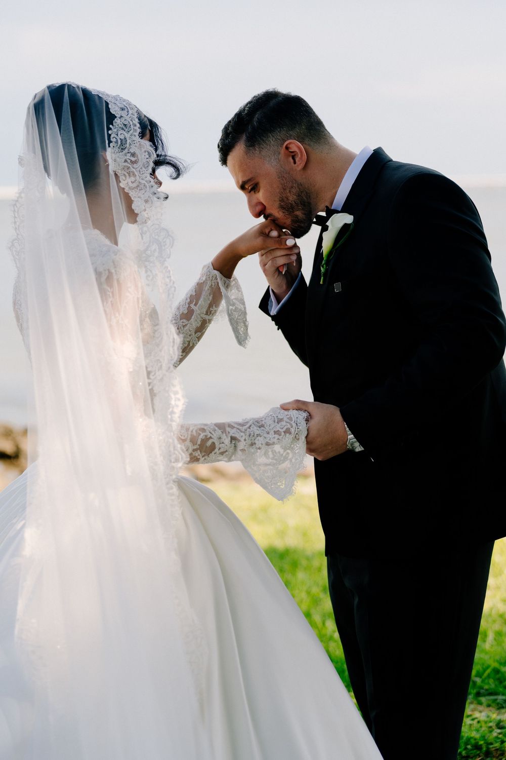 Groom in black suit kissing bride's hand during outdoor wedding ceremony, bride wearing lace veil and gown.