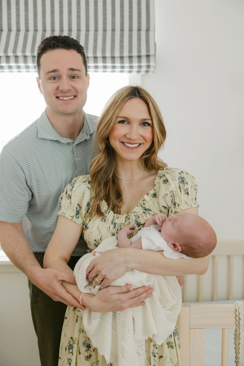 Smiling couple in a bright nursery holding their newborn baby, woman in floral dress, man in green polo shirt.