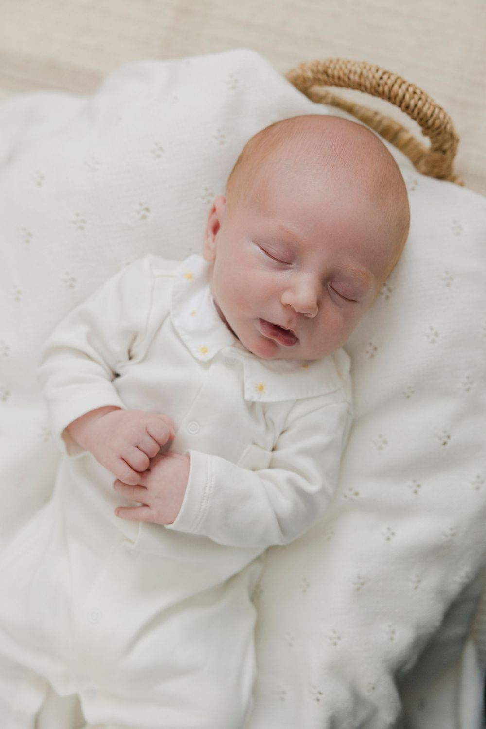 Peaceful newborn baby sleeping in a wicker bassinet, dressed in a white knit romper on a soft blanket.