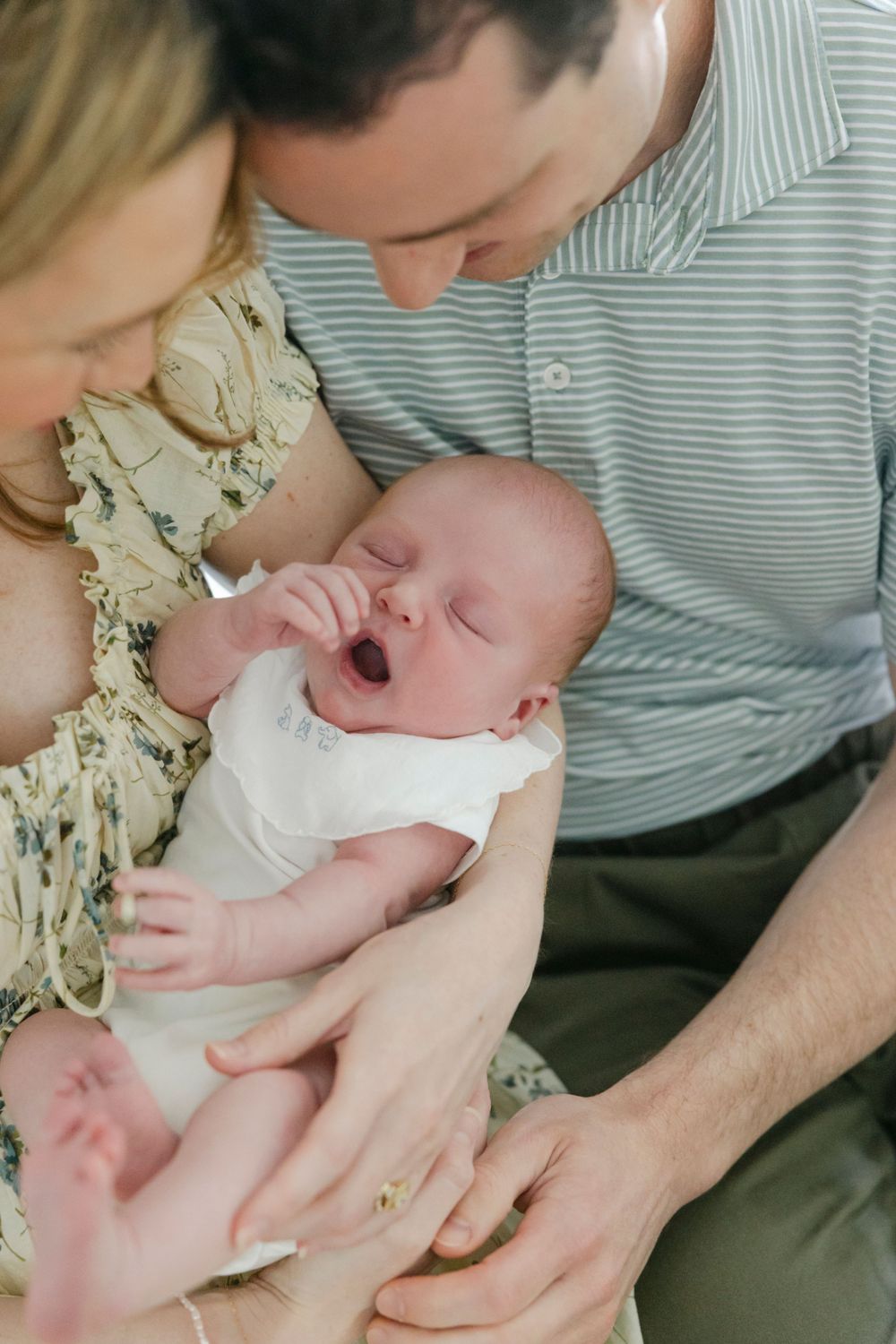New parents lovingly cradle their yawning newborn baby dressed in white, sharing a tender family moment.