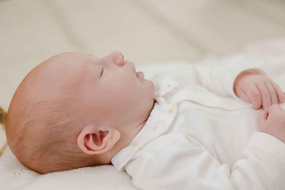 Peaceful newborn baby sleeping on white blanket wearing white onesie, soft natural light nursery photography.
