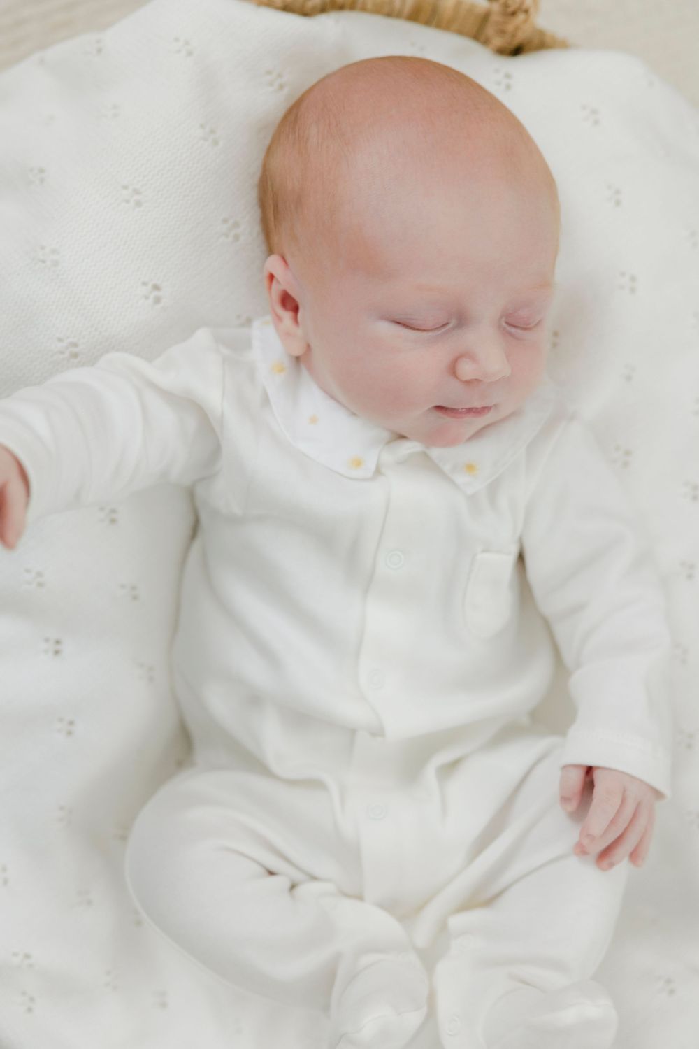 Newborn baby sleeping peacefully in white footed pajamas on a soft white blanket.