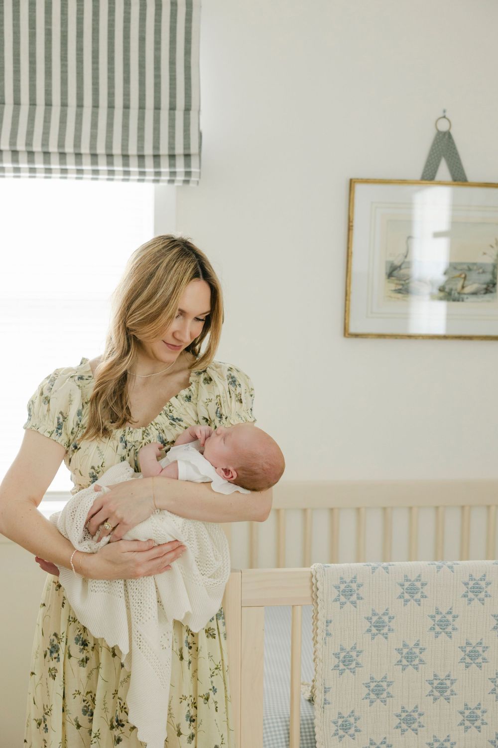 Mother in floral dress tenderly holds newborn baby beside a wooden crib in a soft, naturally lit nursery.