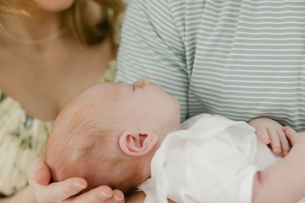 A newborn baby rests peacefully in the arms of two parents, wearing a white outfit in a soft, warm setting.