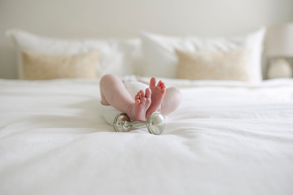 Tiny baby feet peeking out from white bedding on a bright, cozy bed with white pillows in the background.