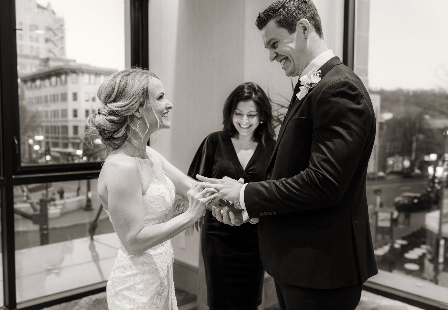 Black and white photo of a bride and groom exchanging rings at their wedding ceremony with an officiant smiling behind them.