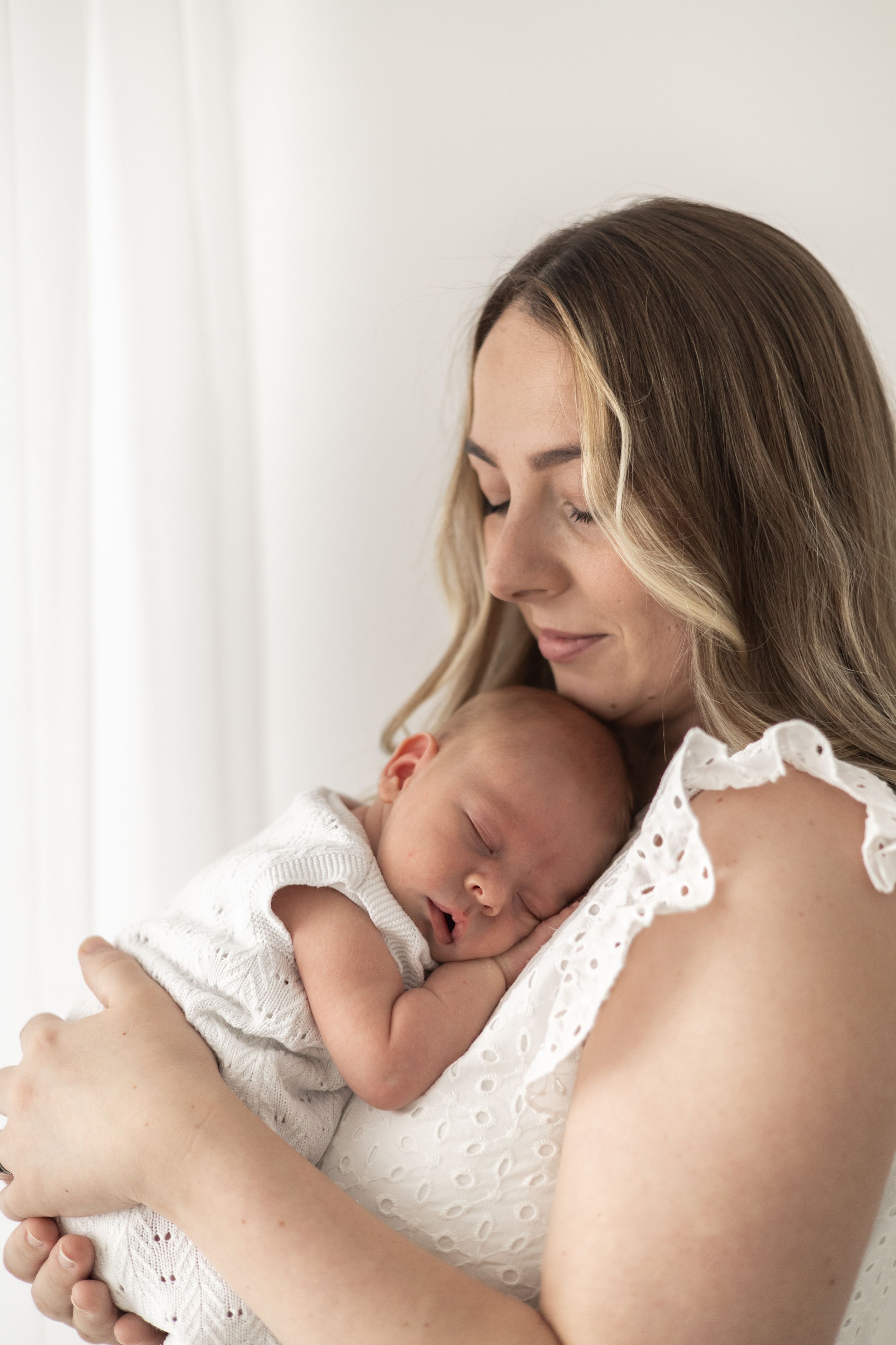 A mother tenderly holds her sleeping newborn baby wrapped in white, relaxed newborn photoshoot Nottingham