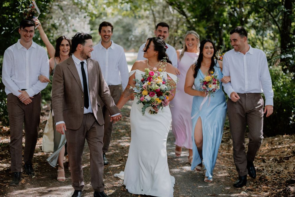 A wedding party walks and laughs together along a tree-lined path on a sunny day.