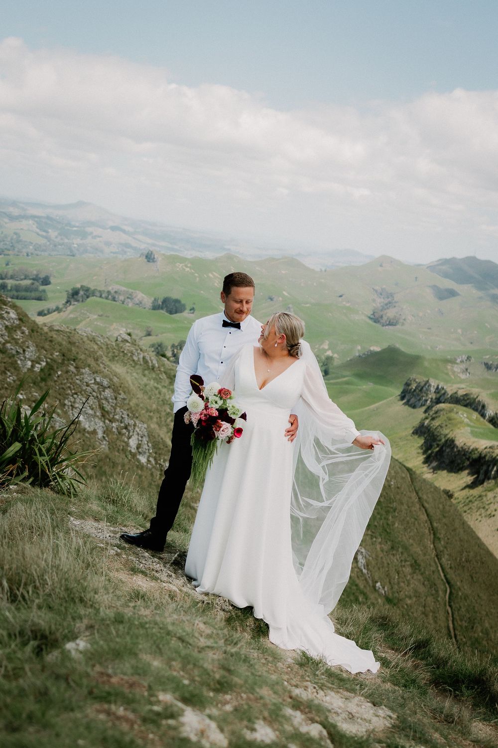 Wedding couple poses on scenic mountain ridge with rolling green hills and dramatic landscape views in background.