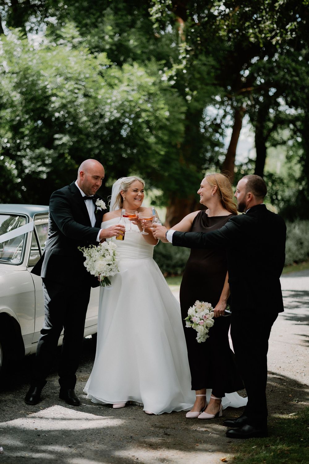 A wedding party in formal attire gathers outdoors near a vintage car on a summer day.
