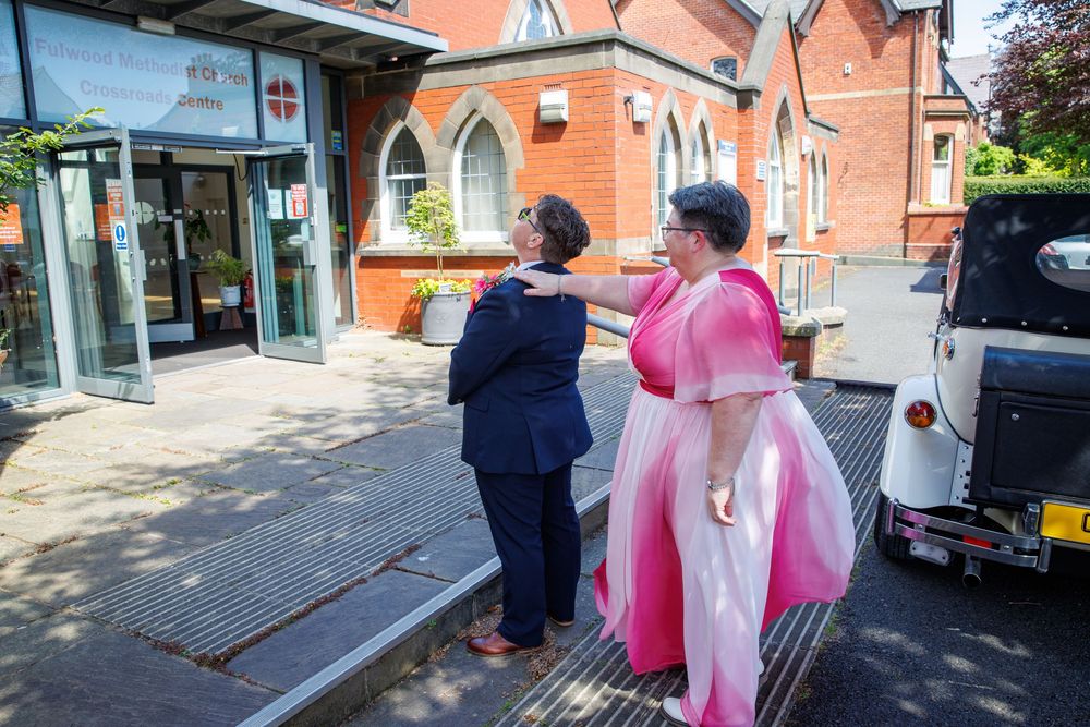 Two women in elegant attire, one in a pink gown, greet outside a red brick church building on a sunny day.