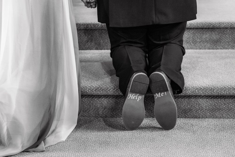 Black and white wedding photo showing groom kneeling with Help Me written on his shoe soles beside bride's dress.