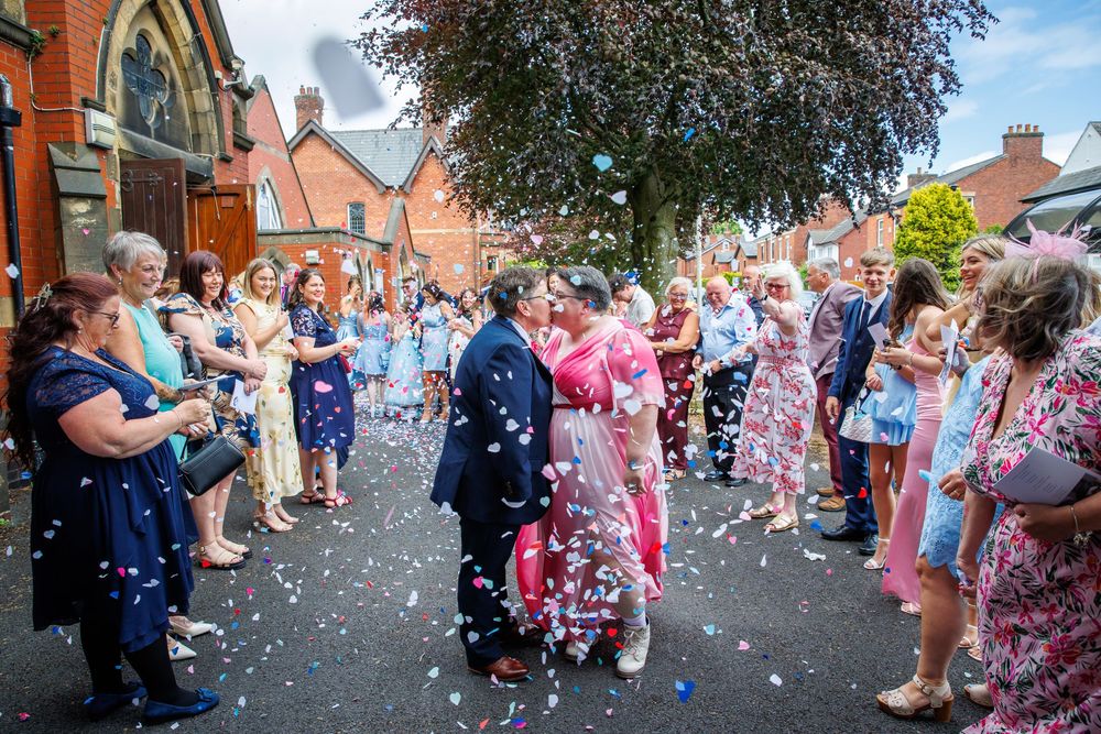 Newlywed couple kissing amid colorful confetti shower, surrounded by cheering wedding guests outside a historic brick church.